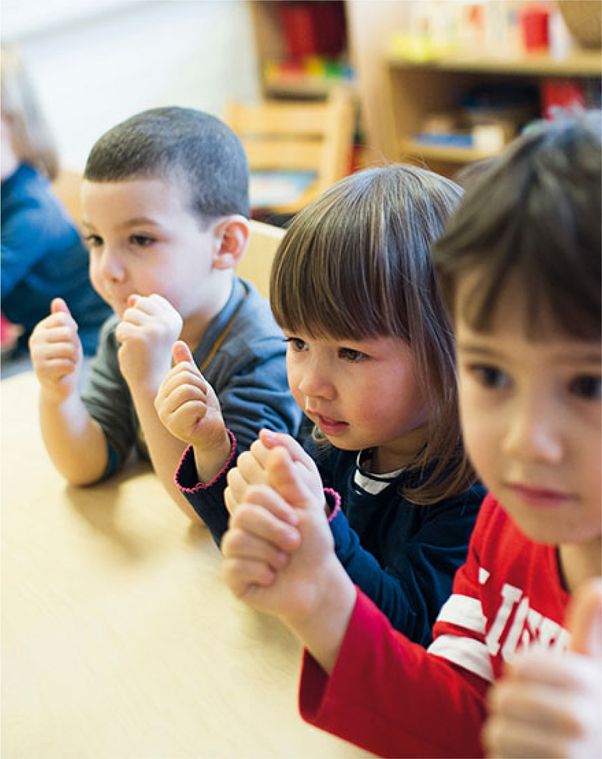 3 kinderen zitten aan tafel. Met ellebogen op tafel en vuisten omhoog